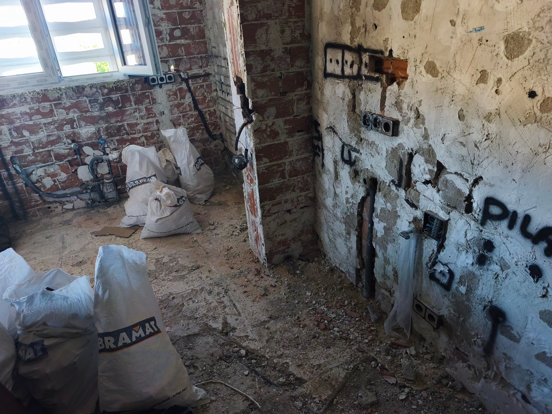 Baño en obra con los azulejos quitados, tomas de agua en la pared y sacos de escombro