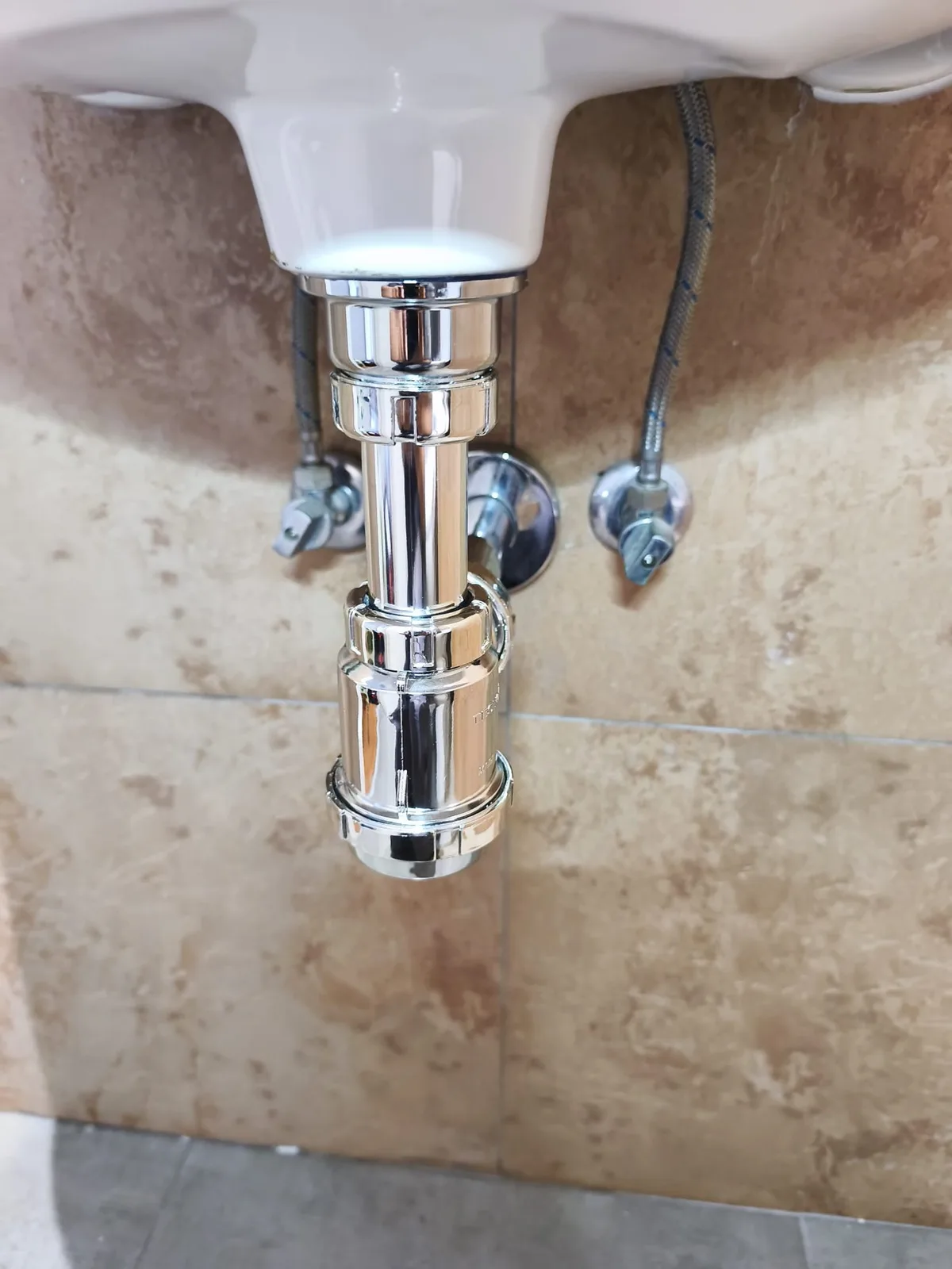 Sifón de botella cromado bajo un lavabo, con sus llaves de escuadra y latiguillos